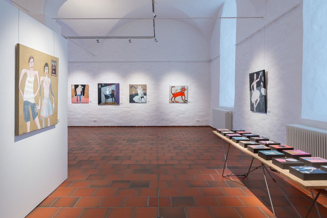 Blick in Ausstellungsraum mit Gemälden an den Wänden und kleinen Arbeiten auf einem Tapeziertisch drapiert