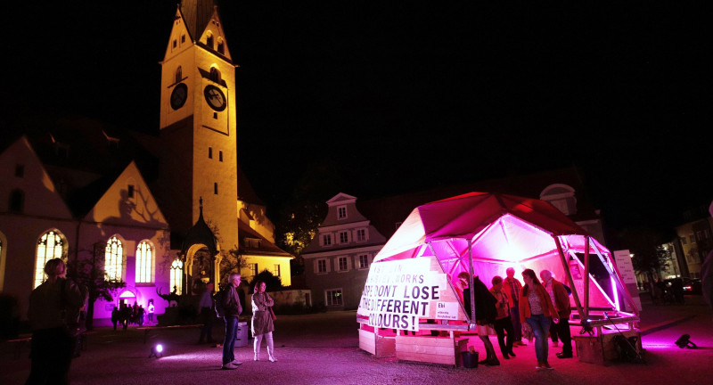 ein pink angeleuchtetes Tipi und eine gelb angeleuchtete Kirche auf einem nächtlichen Platz